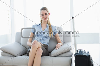 Classy blonde businesswoman waiting sitting on couch next to her suitcase