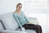 Cute blonde woman holding a book and a cup while sitting on couch