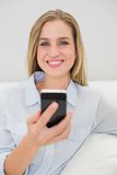 Cheerful casual blonde relaxing on couch holding smartphone