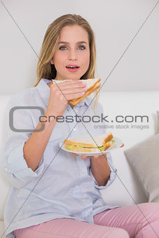 Content casual blonde sitting on couch holding sandwich