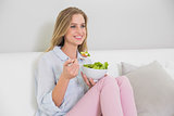 Happy casual blonde sitting on couch holding salad bowl