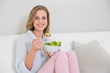 Smiling casual blonde sitting on couch holding salad bowl