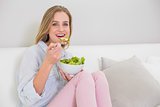 Smiling casual blonde sitting on couch eating salad