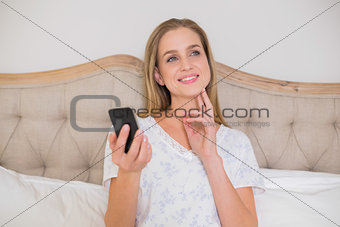 Natural thoughtful woman sitting in bed holding smartphone