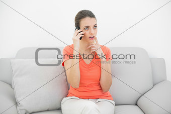 Pretty thinking woman phoning with smartphone sitting on couch