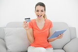 Cheering brunette woman showing her credit card holding her tablet