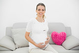 Cute pregnant woman smiling proudly at camera sitting on couch