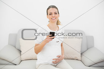 Cute pregnant woman holding her smartphone smiling happily at camera