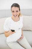 Cheerful pregnant woman holding her smartphone sitting on couch