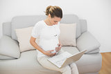 Content ponytailed pregnant woman touching her belly using her notebook