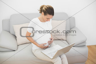 Content ponytailed pregnant woman touching her belly using her notebook