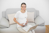 Smiling pregnant woman sitting on couch using her laptop touching her belly