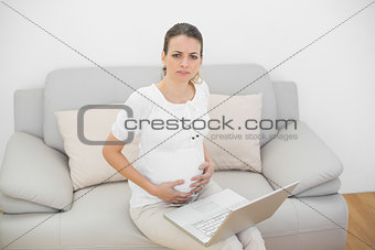 Serious pregnant woman holding her belly sitting on couch