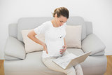 Serious pregnant woman touching her injured back using her notebook