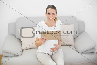 Young cute pregnant woman touching her belly while holding her tablet