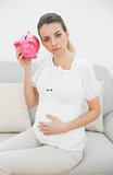 Unhappy pregnant woman shaking a piggy bank while touching her belly