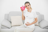 Disappointed pregnant woman shaking a pink piggy bank while holding her belly