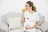 Lovely pregnant woman eating green apple while touching her belly