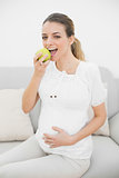 Content pregnant woman eating green apple sitting on couch