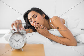 Cute yawning woman lying in her bed while turning off the alarm clock