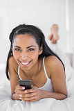 Beautiful smiling woman holding her smartphone lying on her bed