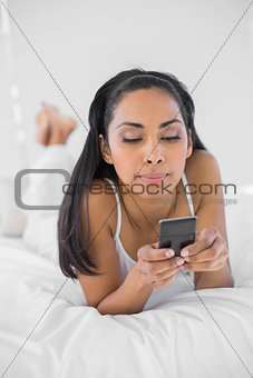 Pretty serious woman using her smartphone lying on her bed