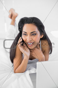 Gleeful woman lying on bed phoning and using her notebook