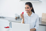 Cute smiling woman holding a strawberry standing in bright kitchen