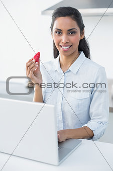Calm woman holding a strawberry using her laptop