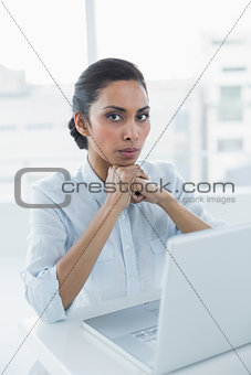 Serious dark haired woman looking at camera sitting at her desk
