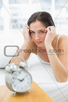 Serious woman with alarm clock lying in bed
