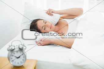 Woman covering ears with pillow and alarm clock on side table