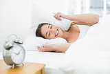 Smiling woman covering ears with pillow and alarm clock on side table