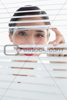 Smiling young business woman peeking through blinds