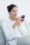 Thoughtful well dressed woman with cellphone and laptop on sofa