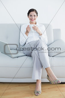 Full length of a smiling well dressed woman with laptop and coffee cup on sofa
