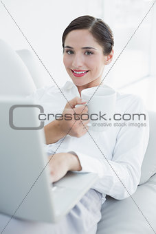 Smiling well dressed woman with laptop and coffee cup on sofa