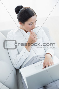 Well dressed woman drinking coffee while using laptop and coffee cup at home