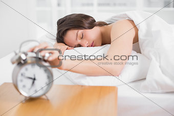 Woman in bed with eyes closed extending hand to alarm clock