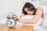 Sleepy woman in bed extending hand to alarm clock
