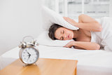 woman covering ears with pillow in bed and alarm clock on side table