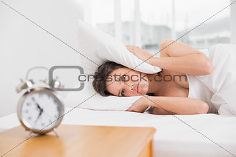 woman covering ears with pillow in bed and alarm clock on table