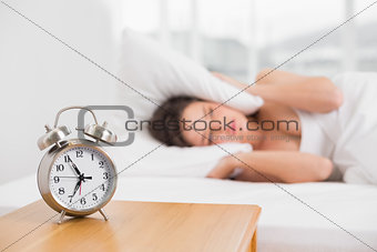 Woman covering ears with pillow in bed and alarm clock on table
