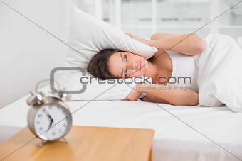 Woman covering ears with pillow in bed and alarm clock on table