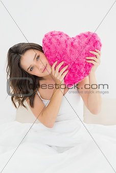 Cute woman holding heart shaped pillow in bed