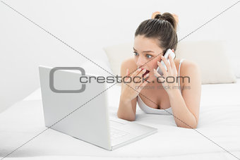 Shocked woman using cellphone and laptop in bed