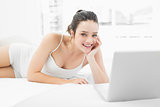 Smiling casual woman with laptop lying in bed