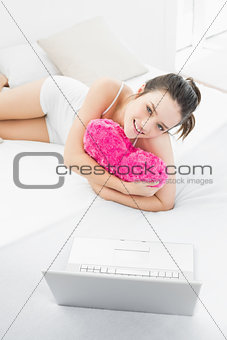 Casual woman with heart shaped pillow and laptop in bed