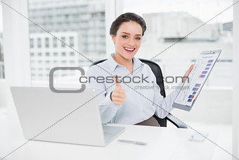 Businesswoman with graphs and laptop gesturing thumbs up in office
