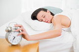 Woman in bed with eyes closed extending hand to alarm clock